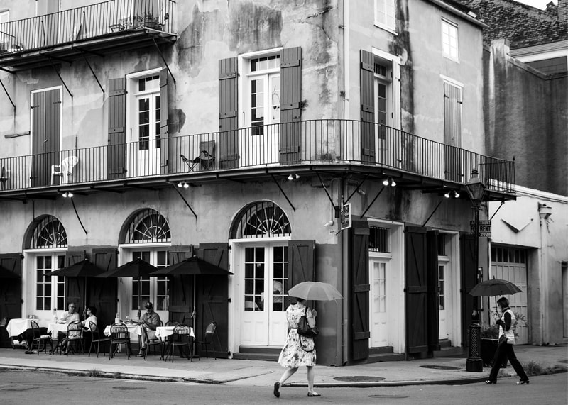 A black and white photograph of two people holding umbrellas and crossing the street in New Orleans iconic French Quarter.